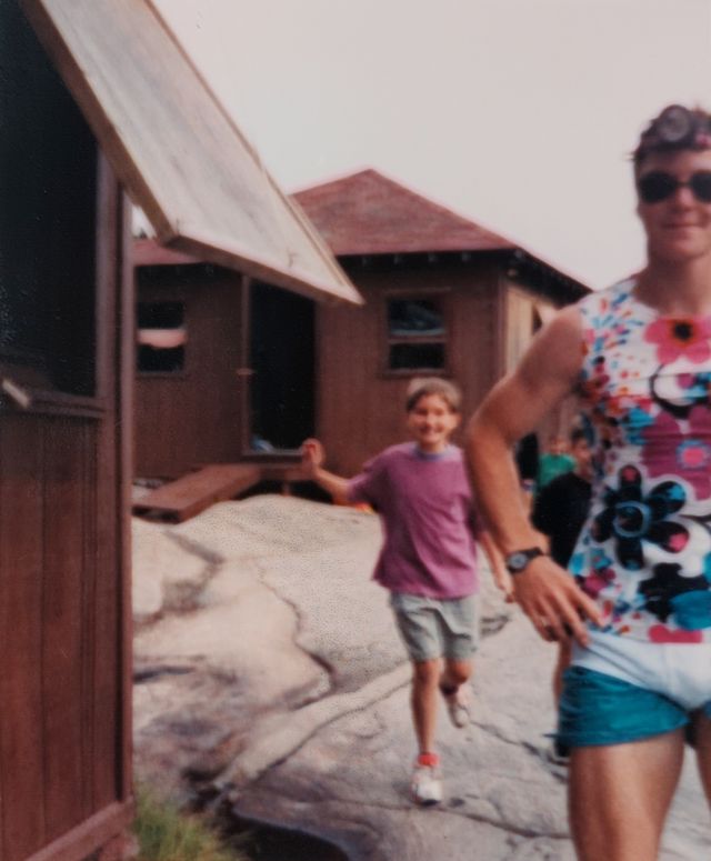 'Cabins with a child in the background and a camp counsellor in the foreground, with his underwear worn outside his shorts.'