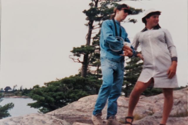 'Two camp counsellors posing on a rocky outcropping, one of them all in denim throwing gang signs, the other in drag, posing with hand on hip.'